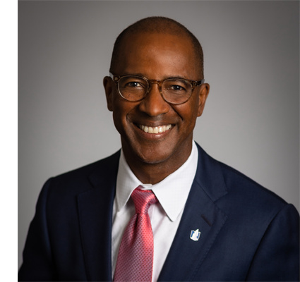 Professional headshot of a smiling man in a navy suit with pink tie and glasses